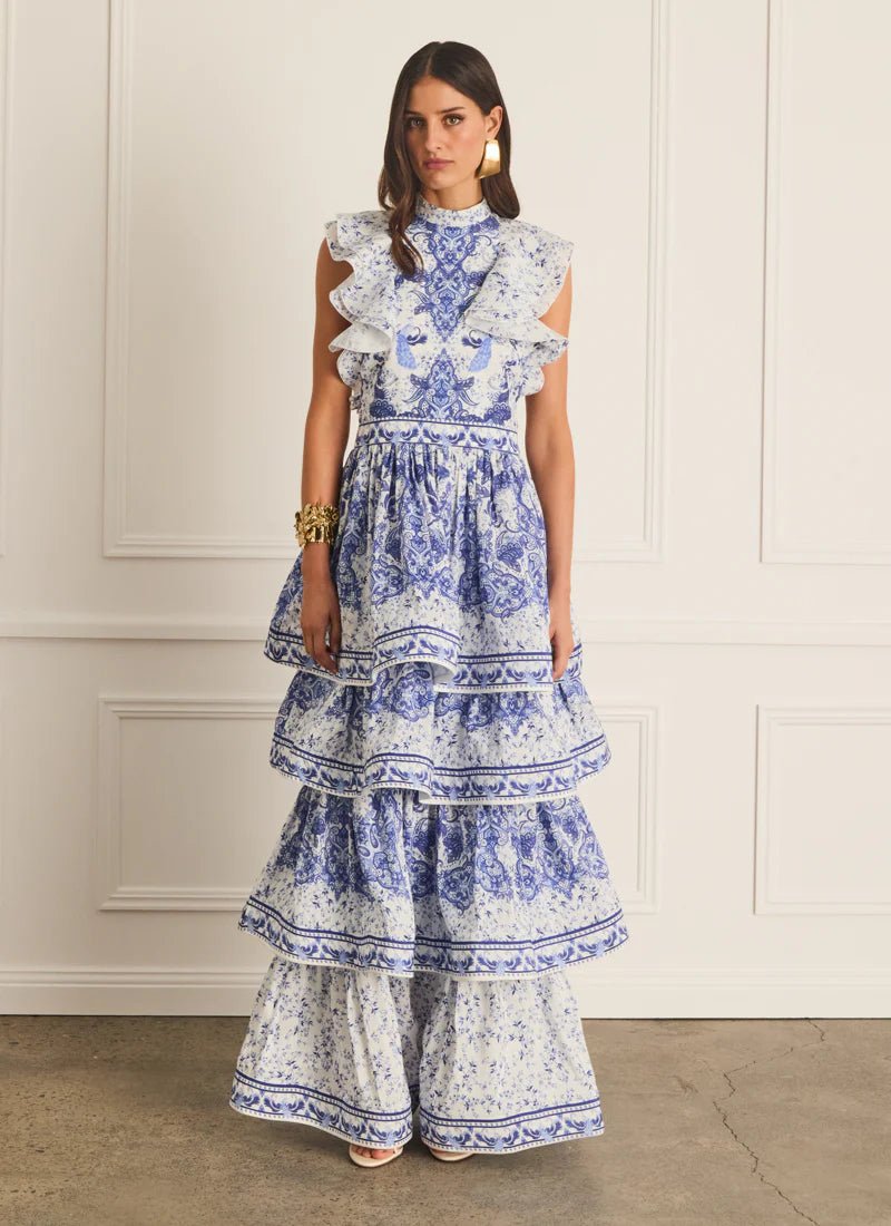 Full length front view of the Aqua Blu Willow Lily Maxi Dress in a blue and white floral porcelain print, styled with gold statement jewelry for a high-end summer event look.