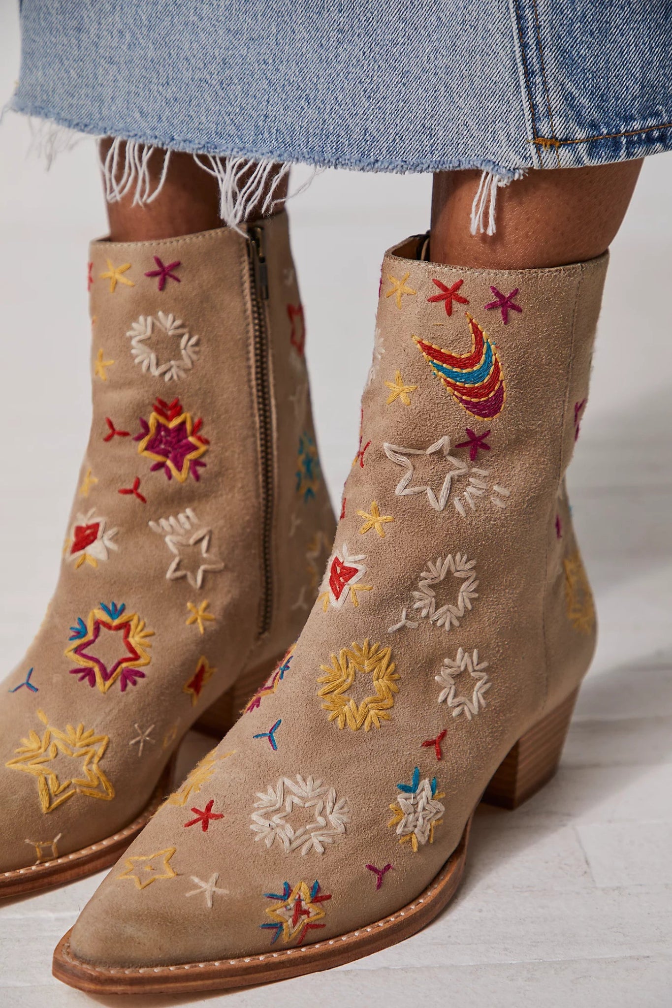 A pair of beige Free People Bowers ankle boots with colorful star and heart embroidery, pointed toe, and stacked block heel, worn with denim pants.