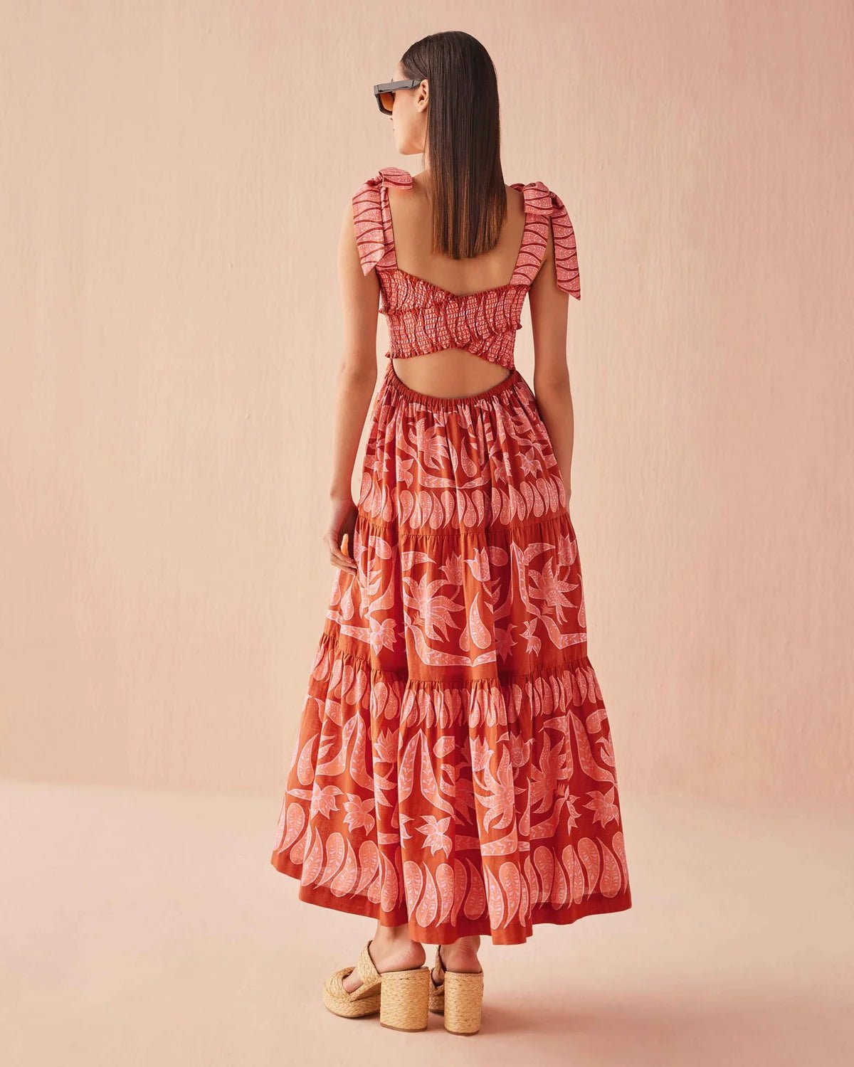Back view of the Omika Lana Maxi Dress in Vivi Negroni print, featuring a vibrant orange and pink tropical floral pattern with an open cross-back cutout and tiered skirt.