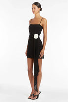 Side view of a woman wearing the black Amanda Uprichard Isabel Mini Dress with white rosette and long drape detail