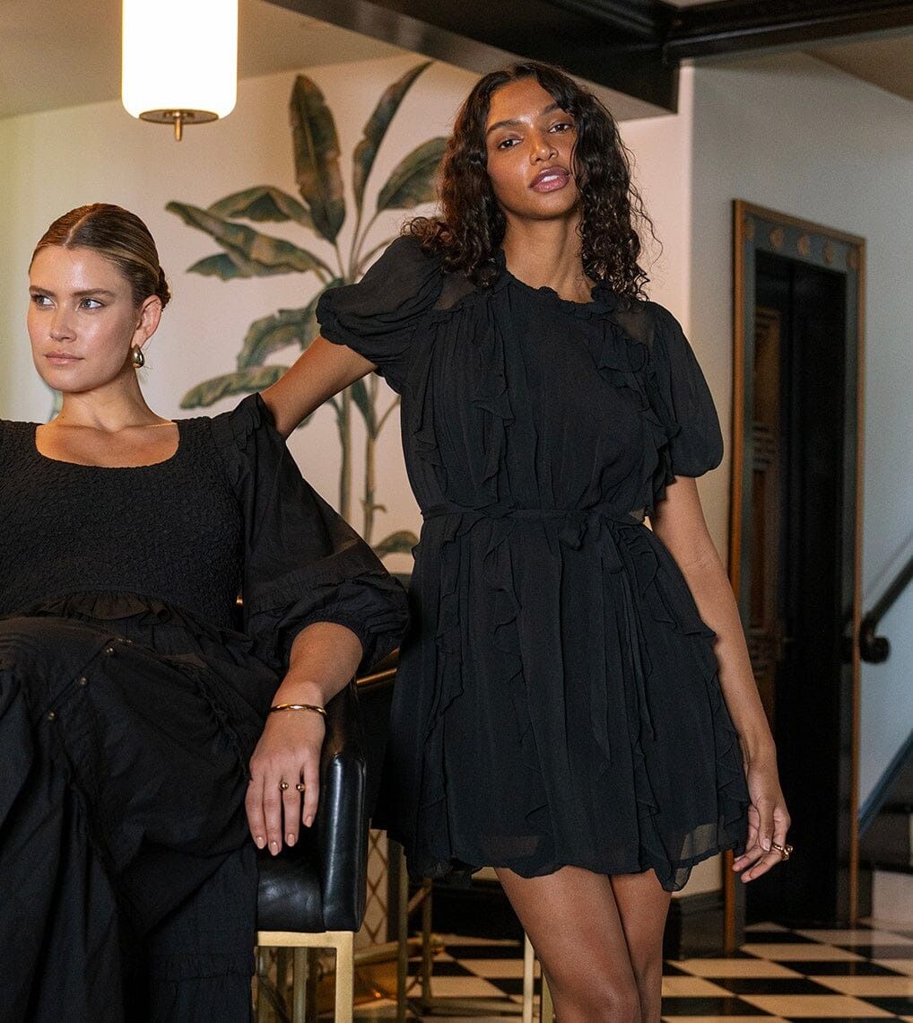 A woman in a black ruffled mini dress with puff sleeves stands next to another woman in a black outfit in an indoor setting with a checkered floor and a potted plant in the background.