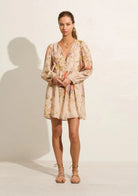 A woman modeling the Auguste the Label mini dress with a colorful floral print, long balloon sleeves, and a V-neckline, standing against a white studio wall with a soft shadow.