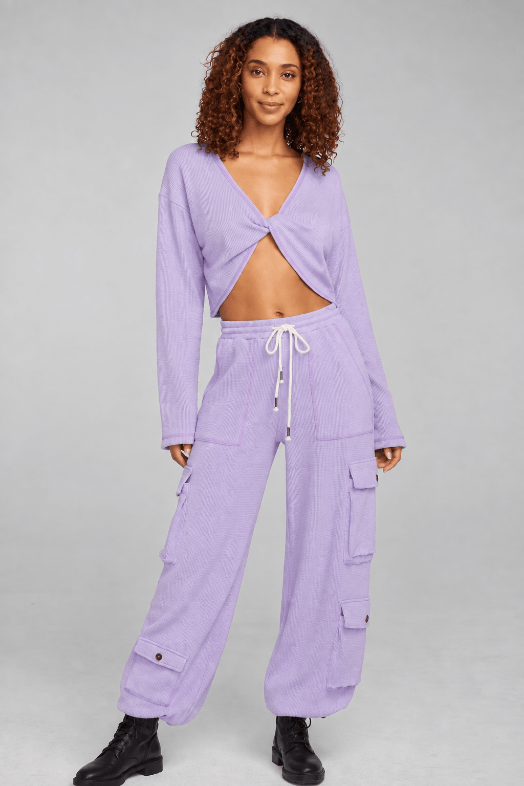 A model wearing a matching lavender two-piece lounge set consisting of a long-sleeve wrap-front crop top and wide-leg cargo pants with side pockets, styled with black boots against a solid gray studio background.