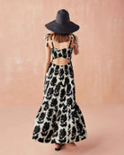 Back view of the Omika Lana Maxi Dress in Emma Mussel print, featuring a stylish open cross-back cutout, self-tie shoulder straps, and a tiered skirt styled with a black sun hat.