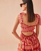 Close-up of the Omika Lana Maxi Dress in Vivi Negroni styled with oversized sunglasses, highlighting the vibrant orange and pink hand-painted print, smocked bodice, and adjustable self-tie shoulder straps.