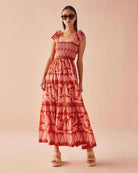 Model wearing the Omika Lana Maxi Dress in Vivi Negroni, a vibrant orange and pink hand-painted floral print on a cotton-linen blend, featuring a smocked bodice and self-tie shoulder straps.